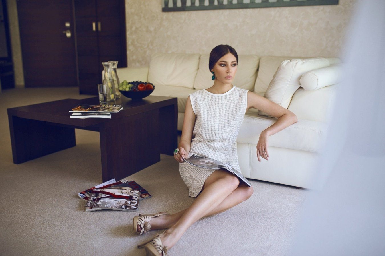 Elegancka kobieta siedzi na podłodze obok białej skórzanej sofy, czytając magazyn w jasnym salonie z ciemnobrązowym stolikiem kawowym i owocami w misce.