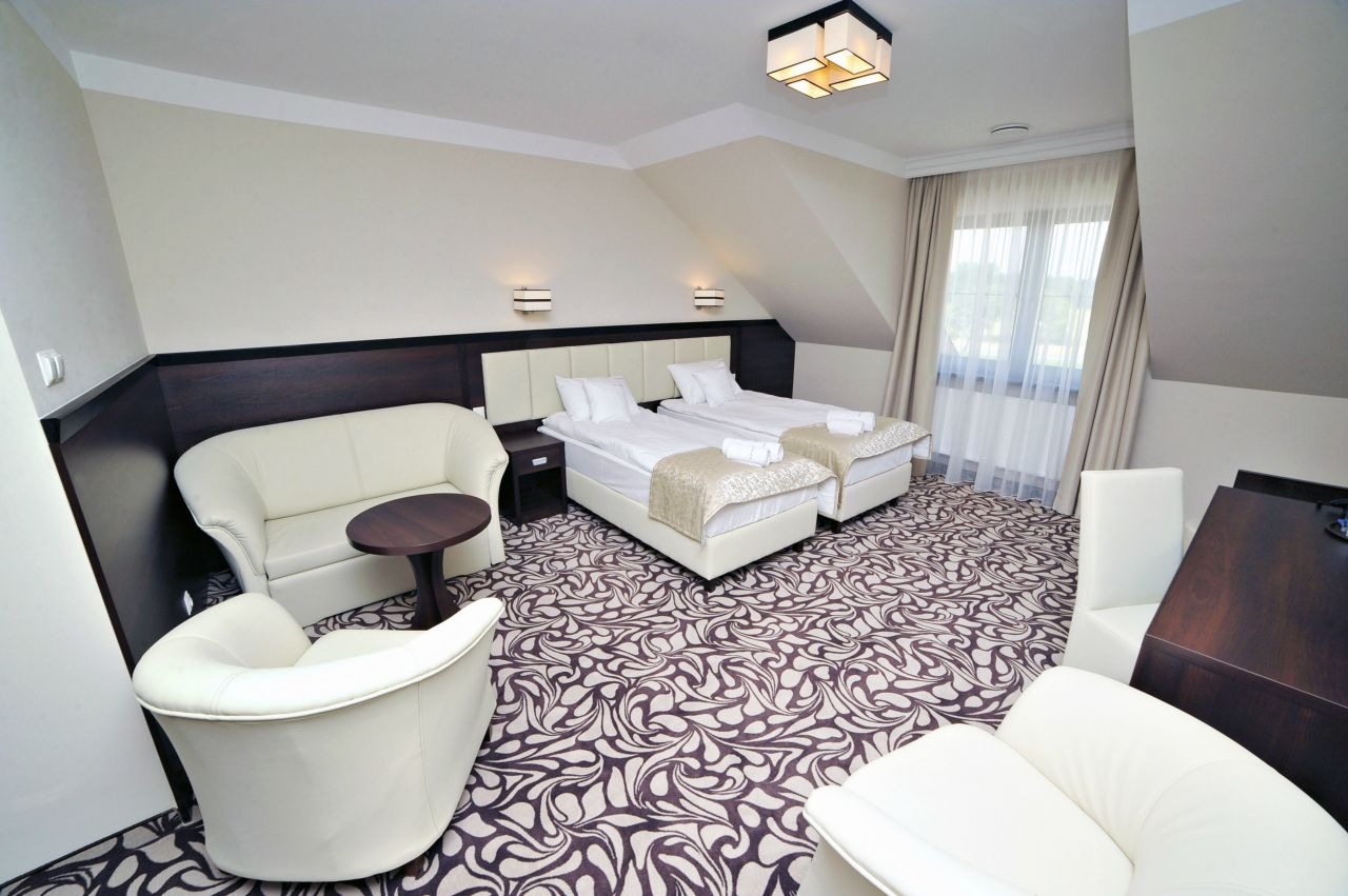 Pokój hotelowy na poddaszu z dwoma łóżkami, sofą, fotelem i wzorzystą wykładziną, oświetlony lampą sufitową i kinkietami.