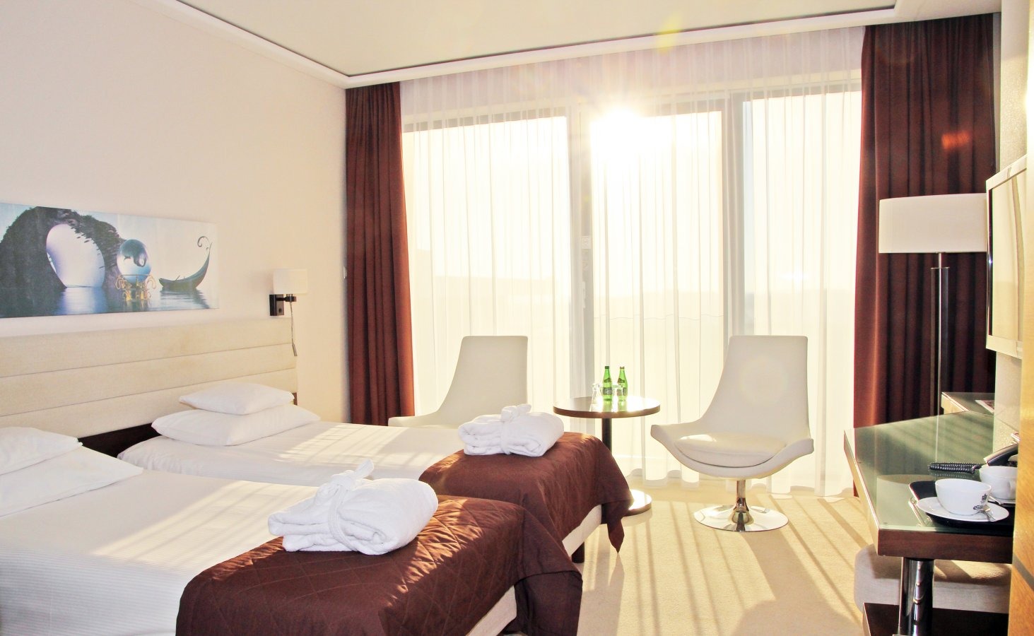 Jasny pokój hotelowy z dwoma łóżkami pojedynczymi, białymi szlafrokami, nowoczesnymi krzesłami i obrazem z motywem morskim na ścianie, oświetlony promieniami słonecznymi wpadającymi przez okno.