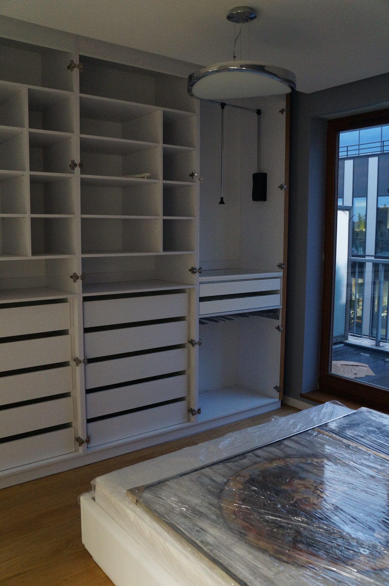 Biała, otwarta szafa z półkami i szufladami, wyposażona w drążek na ubrania, oświetlona lampą sufitową, z widocznym obrazem w folii ochronnej na podłodze.