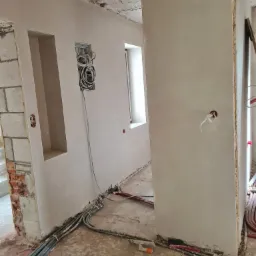 Świeżo otynkowane ściany w surowym wnętrzu, widoczne kable elektryczne i instalacje, ślady po pracach budowlanych na podłodze, przygotowane otwory pod gniazdka i wnęki w ścianach.