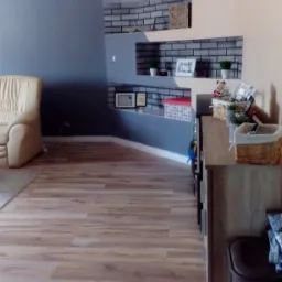 Salon z panelami podłogowymi w odcieniach drewna, szarą ścianą i dekoracyjnymi półkami z cegły, uzupełniony beżowym fotelem i zegarem ściennym.