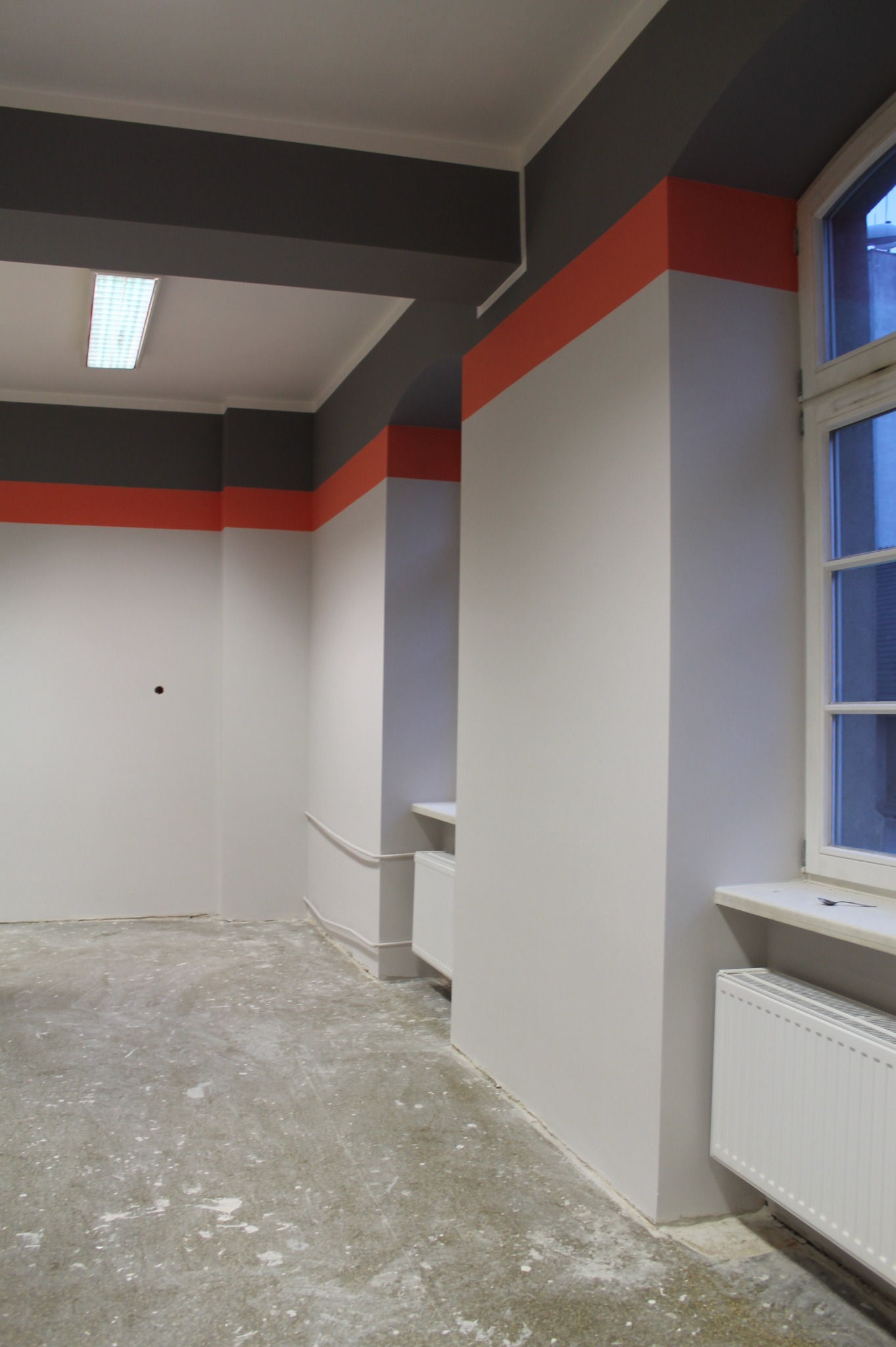 Wnętrze biura po malowaniu ścian na biało z szarymi i pomarańczowymi akcentami, widoczne grzejniki i betonowa podłoga z resztkami farby.