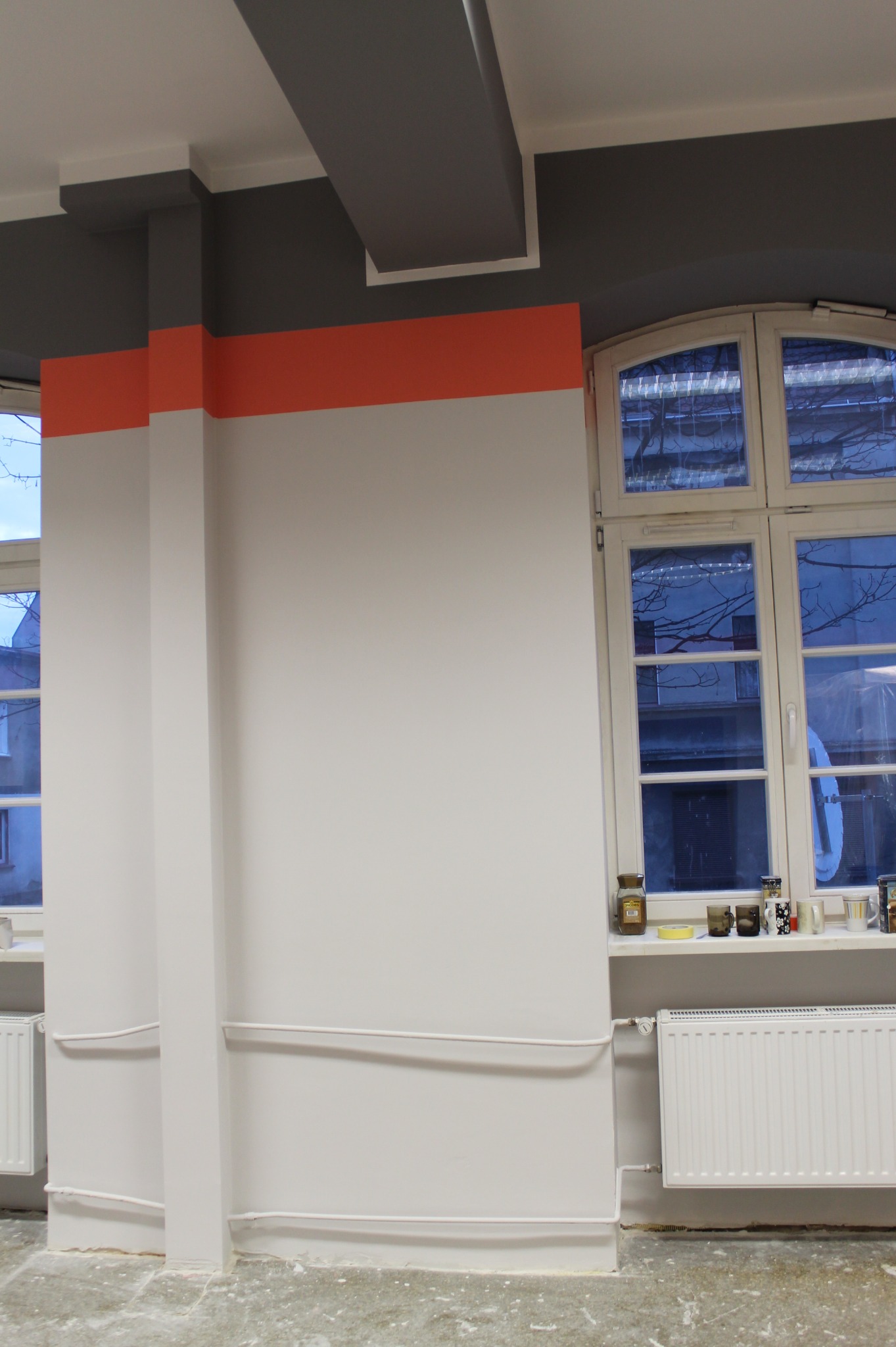 Wnętrze w trakcie malowania: białe ściany z poziomym pomarańczowym pasem, szary sufit, okno z widokiem na drzewa, grzejnik i ślady farby na podłodze.