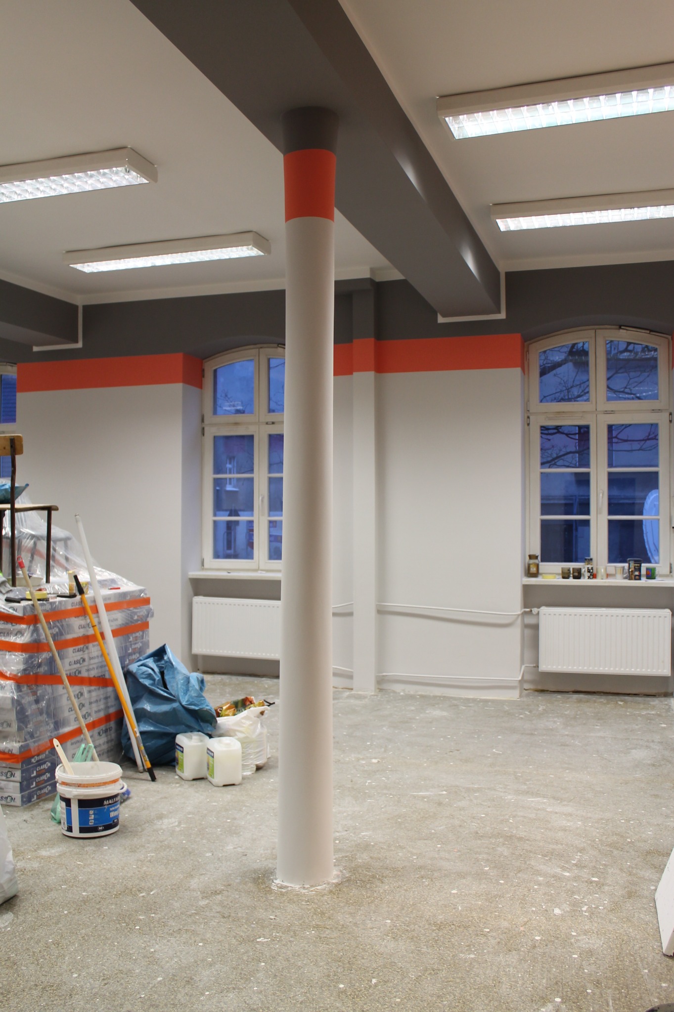 Wnętrze biura w trakcie malowania: białe ściany z szarym pasem pod sufitem i pomarańczowym akcentem, biała kolumna z pomarańczowym paskiem, okna, grzejniki, widoczne narzędzia malarskie i farby...