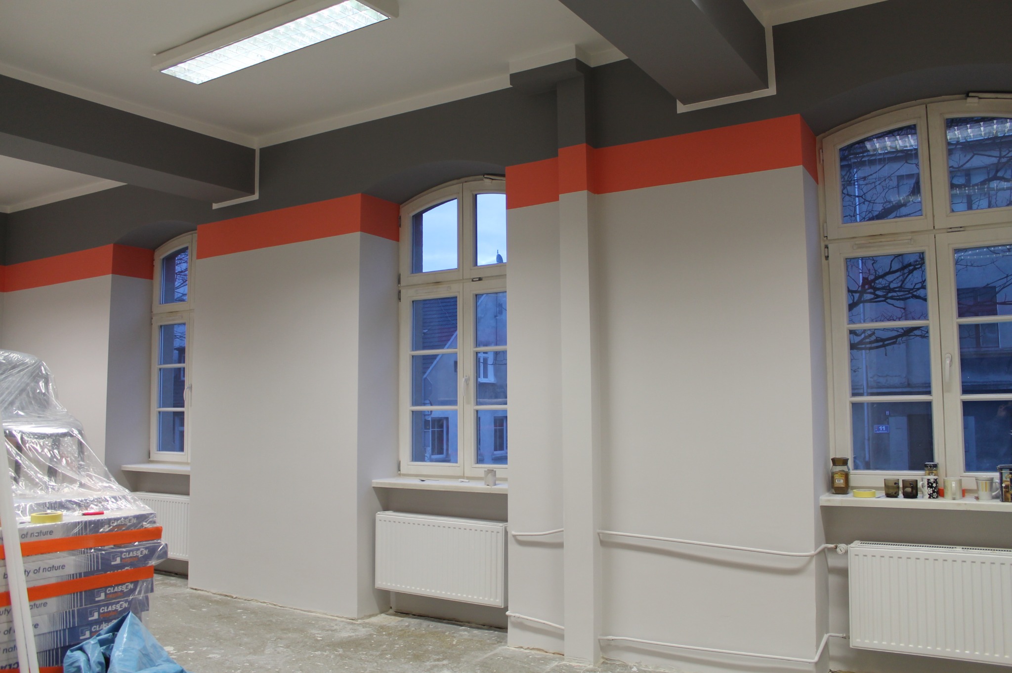 Wnętrze biura po malowaniu: ściany pomalowane na biało z szarym pasem u góry i pomarańczowym akcentem, widoczne trzy okna z białymi ramami, grzejniki, podłoga zabezpieczona folią, w rogu stos...