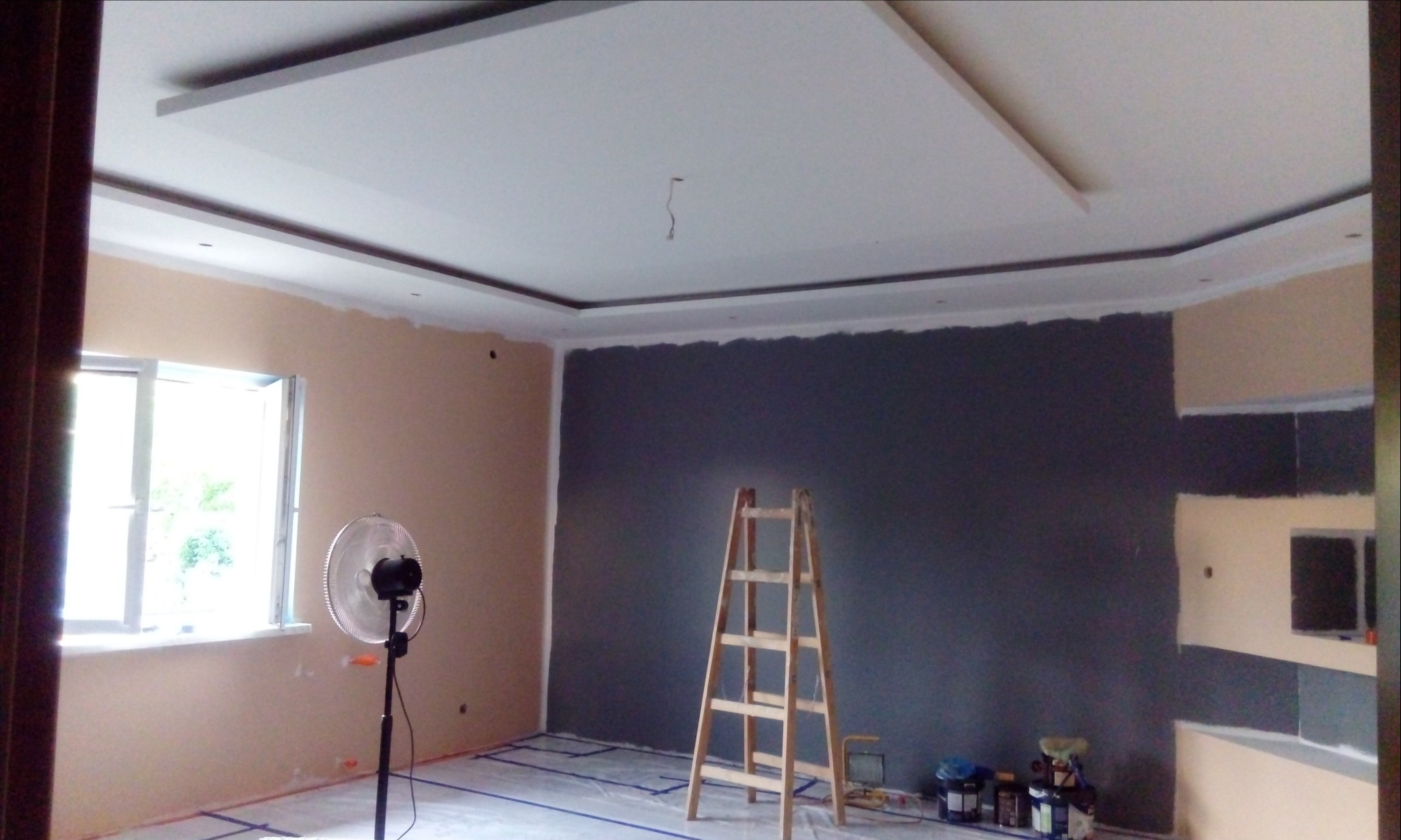 Pokój w trakcie malowania: jedna ściana w kolorze grafitowym, druga beżowa, drewniana drabina, wentylator stojący przy oknie, zabezpieczona folią podłoga.