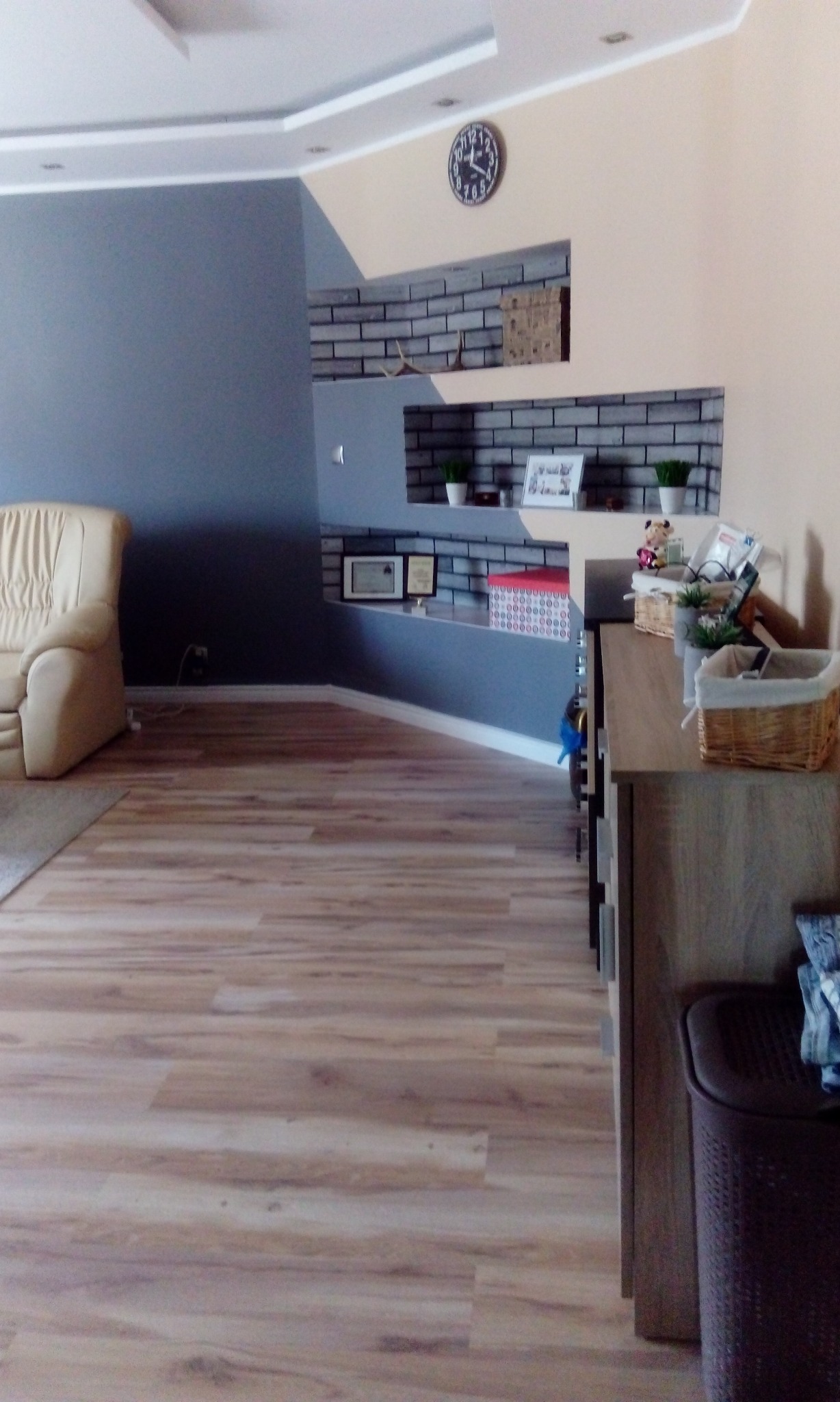 Salon z panelami podłogowymi w odcieniach drewna, szarą ścianą i dekoracyjnymi półkami z cegły, uzupełniony beżowym fotelem i zegarem ściennym.