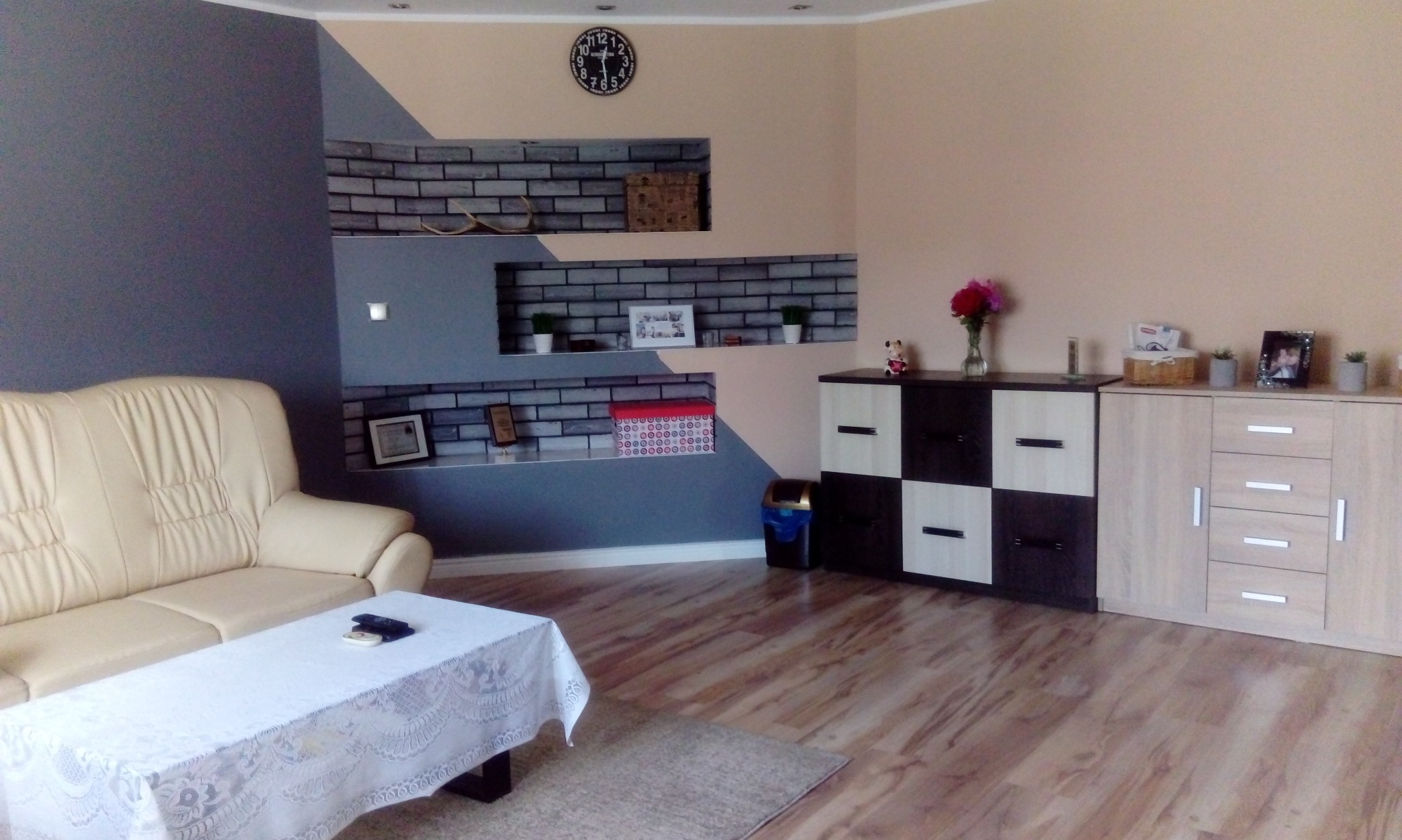 Salon po odświeżeniu z kremową sofą, stolikiem przykrytym białą koronkową serwetą, ścianą pomalowaną w geometryczne wzory w odcieniach szarości i beżu oraz wnękami wyłożonymi imitacją cegły.