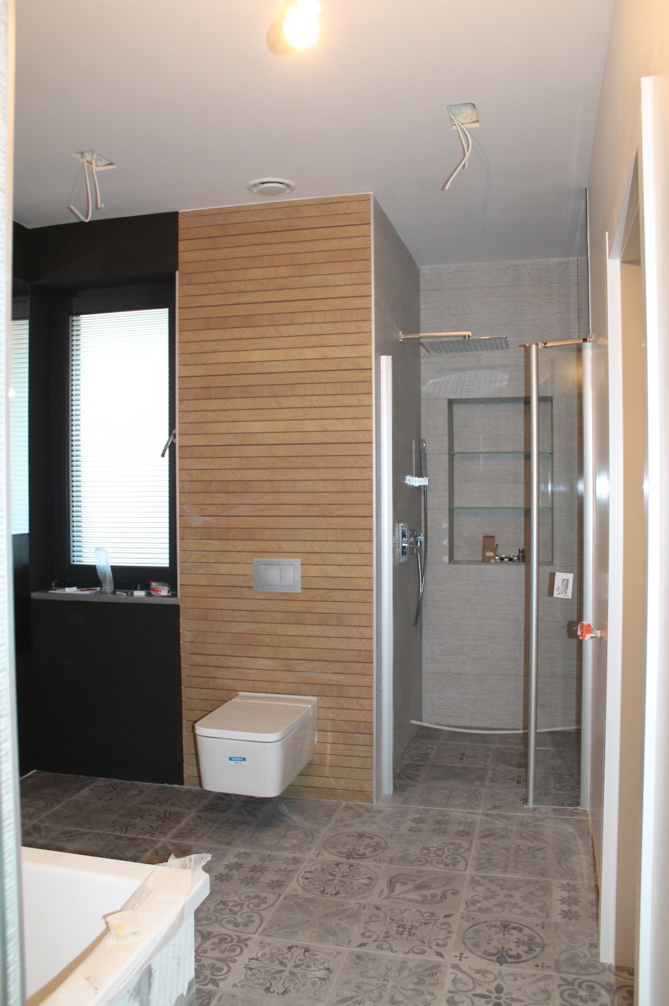 Nowoczesna łazienka w trakcie wykańczania: drewniana boazeria, podwieszana toaleta, kabina prysznicowa typu walk-in z szarymi płytkami, wzorzysta podłoga, niezabezpieczone kable oświetleniowe.