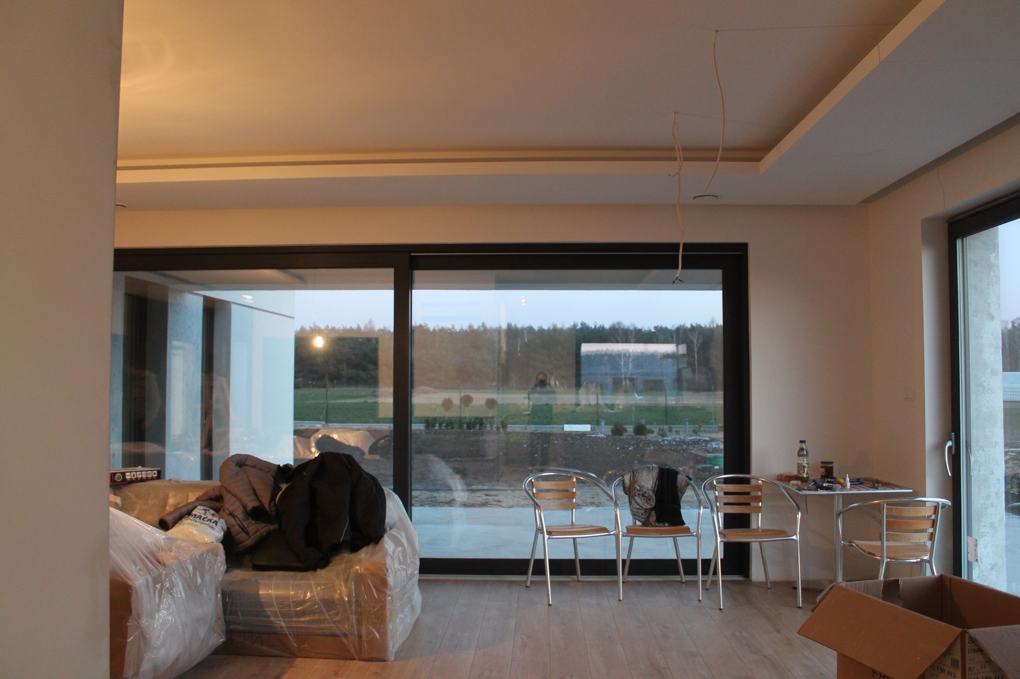 Wnętrze pokoju z dużym oknem przesuwnym, podłogą z paneli, meblami w folii ochronnej, krzesłami i stolikiem z narzędziami, sugerujące prace wykończeniowe.