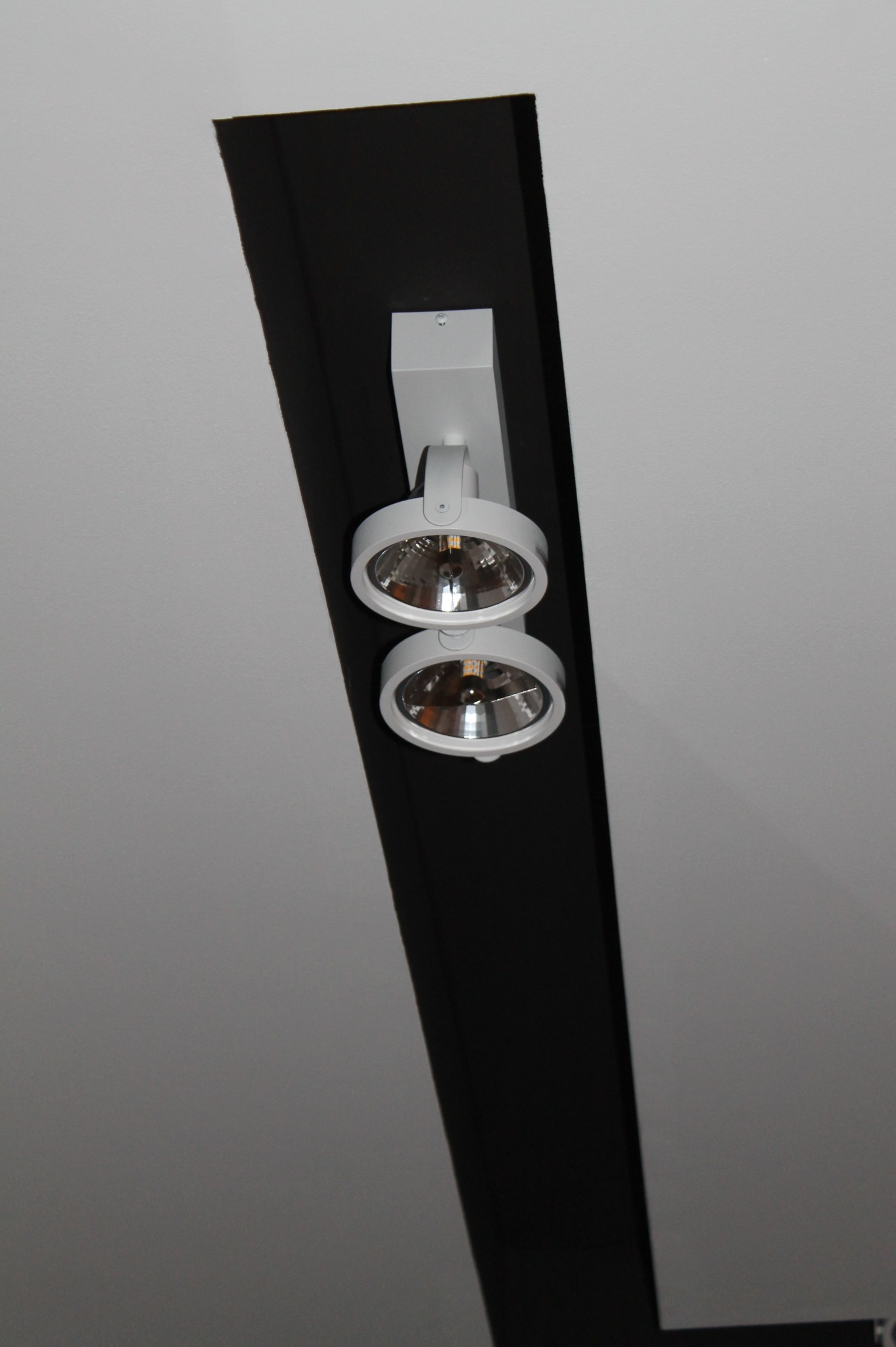 Biała, podwójna lampa reflektorowa zamontowana na czarnej belce sufitowej, oświetlająca biały sufit.