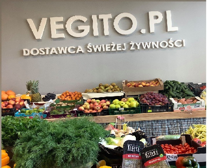 Stoisko z różnorodnymi świeżymi owocami i warzywami, w tle logo 'VEGITO.PL - DOSTAWCA ŚWIEŻEJ ŻYWNOŚCI' na szarej ścianie.