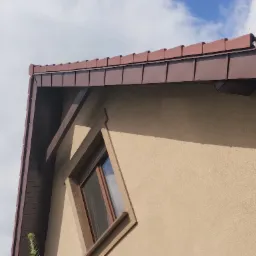 Fragment brązowego dachu z ceramiczną dachówką i obróbką blacharską deski szczytowej, widoczny narożnik dachu i okno w ścianie budynku o beżowej elewacji na tle pochmurnego nieba.