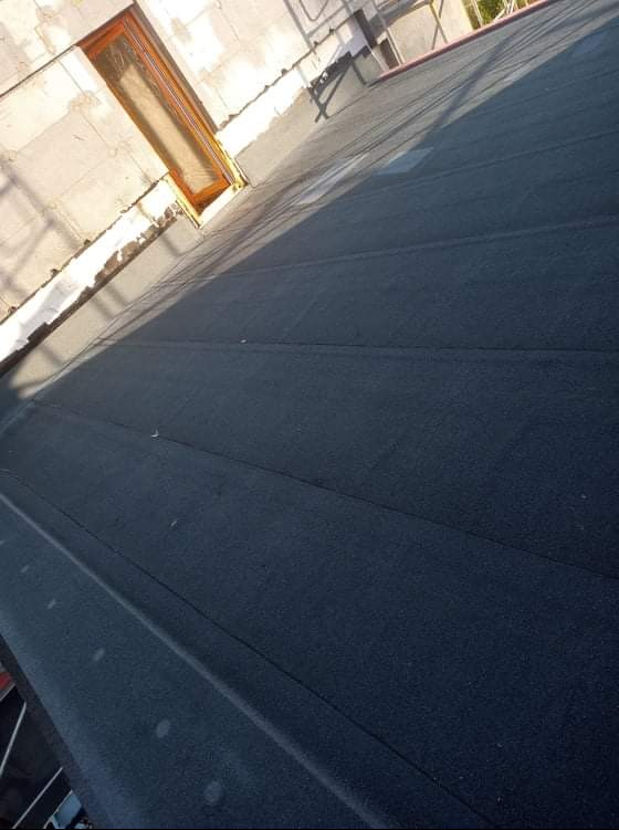 Czarny, płaski dach pokryty papą termozgrzewalną, widoczny fragment okna dachowego i ściana budynku z pustaków.