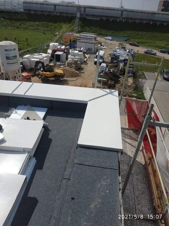 Widok z góry na fragment dachu z szarą papą i białe, prostokątne elementy wykończeniowe, w tle plac budowy z żółtą koparką i zbiornikiem na materiały.