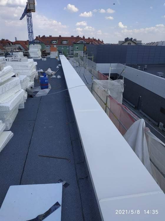 Biała, długa obróbka blacharska zamontowana na dachu pokrytym papą, w tle styropian i budynki miejskie oraz żuraw budowlany.