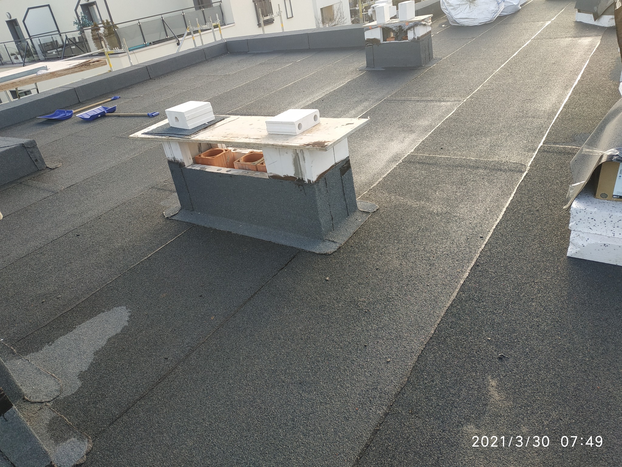 Widok z góry na płaski dach pokryty papą termozgrzewalną z wykończonym kominem i narzędziami dekarskimi w tle.