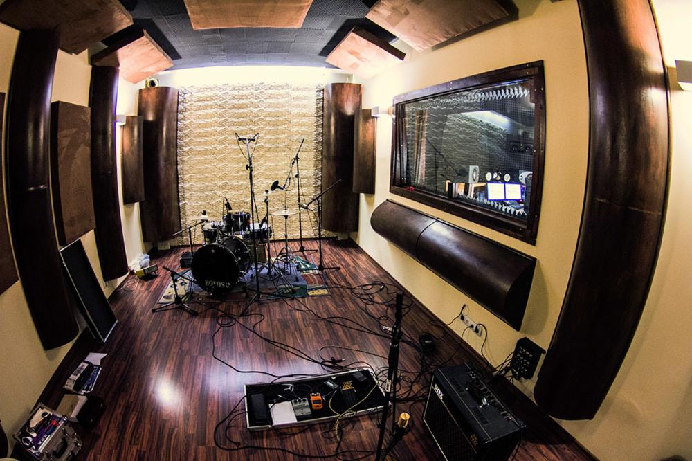 Wnętrze pokoju z perkusją, mikrofonami i wzmacniaczem, widok przez szerokokątny obiektyw, z wygłuszonymi ścianami i sufitem. Okno odsłuchowe z widokiem na sąsiednie pomieszczenie.