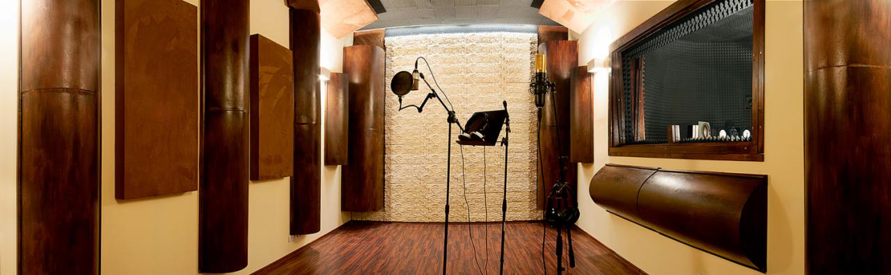 Wnętrze studia nagrań z mikrofonami, panelami akustycznymi i oknem odsłuchowym, oświetlone ciepłym światłem.