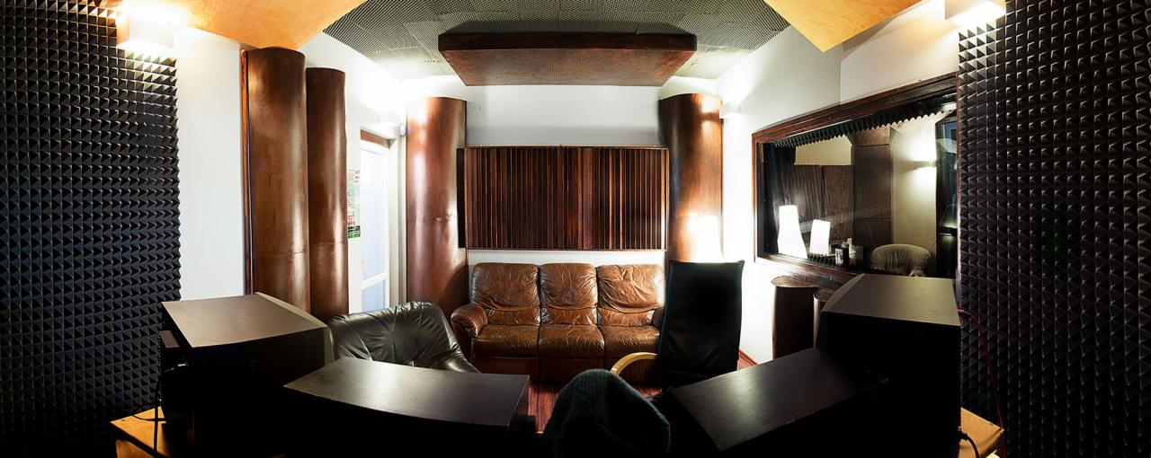 Wnętrze studia nagrań z panelami akustycznymi na ścianach, brązowymi kolumnami i drewnianymi elementami dekoracyjnymi, skórzana sofa i fotele dla komfortu lektorów.