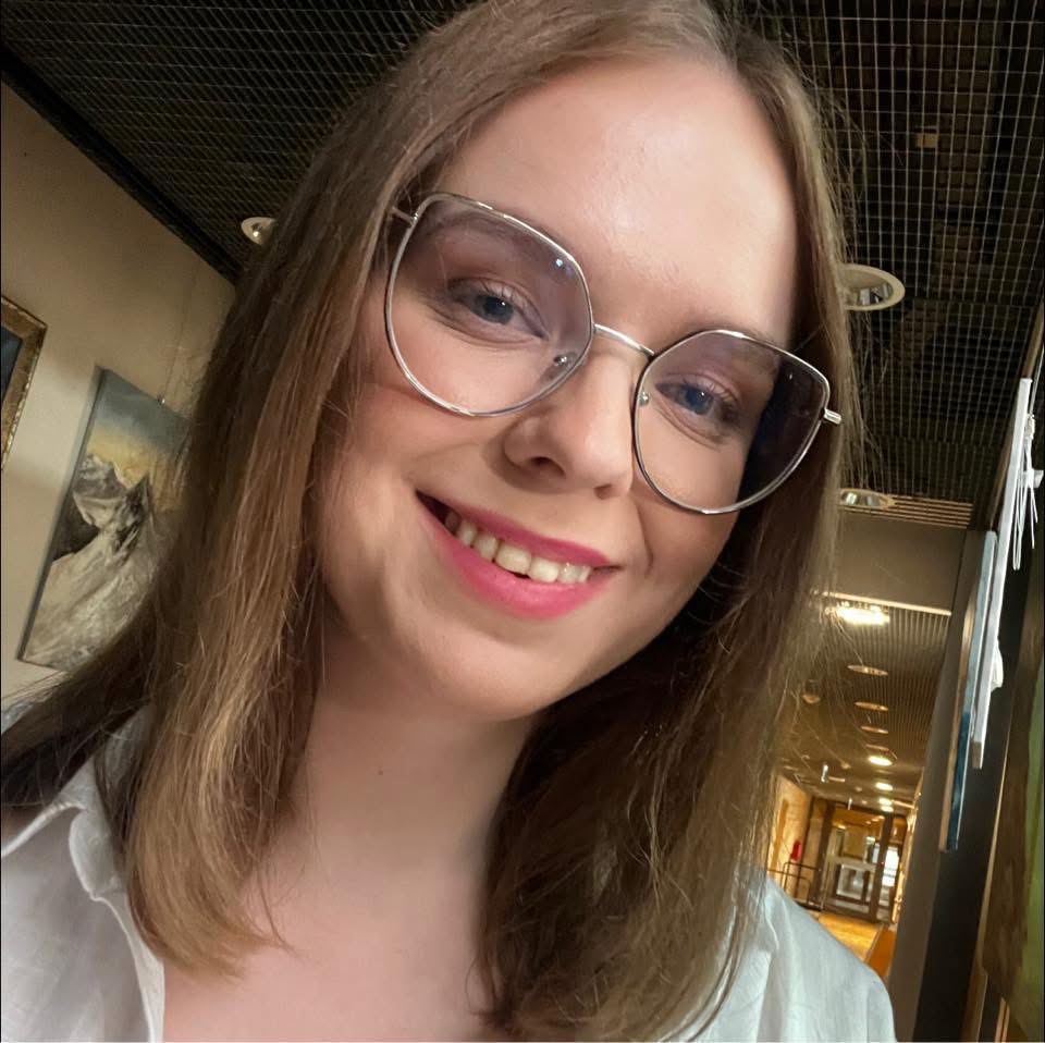 Portret uśmiechniętej kobiety w okularach na tle korytarza z obrazami. Jasne oświetlenie, widoczny sufit z kratką. Ujęcie z bliska, podkreślające detale twarzy.