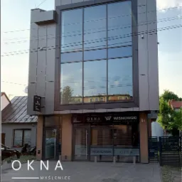 Nowoczesny budynek salonu sprzedaży stolarki okiennej i drzwiowej z dużym przeszkleniem, widok z zewnątrz, w tle zabudowa miejska.