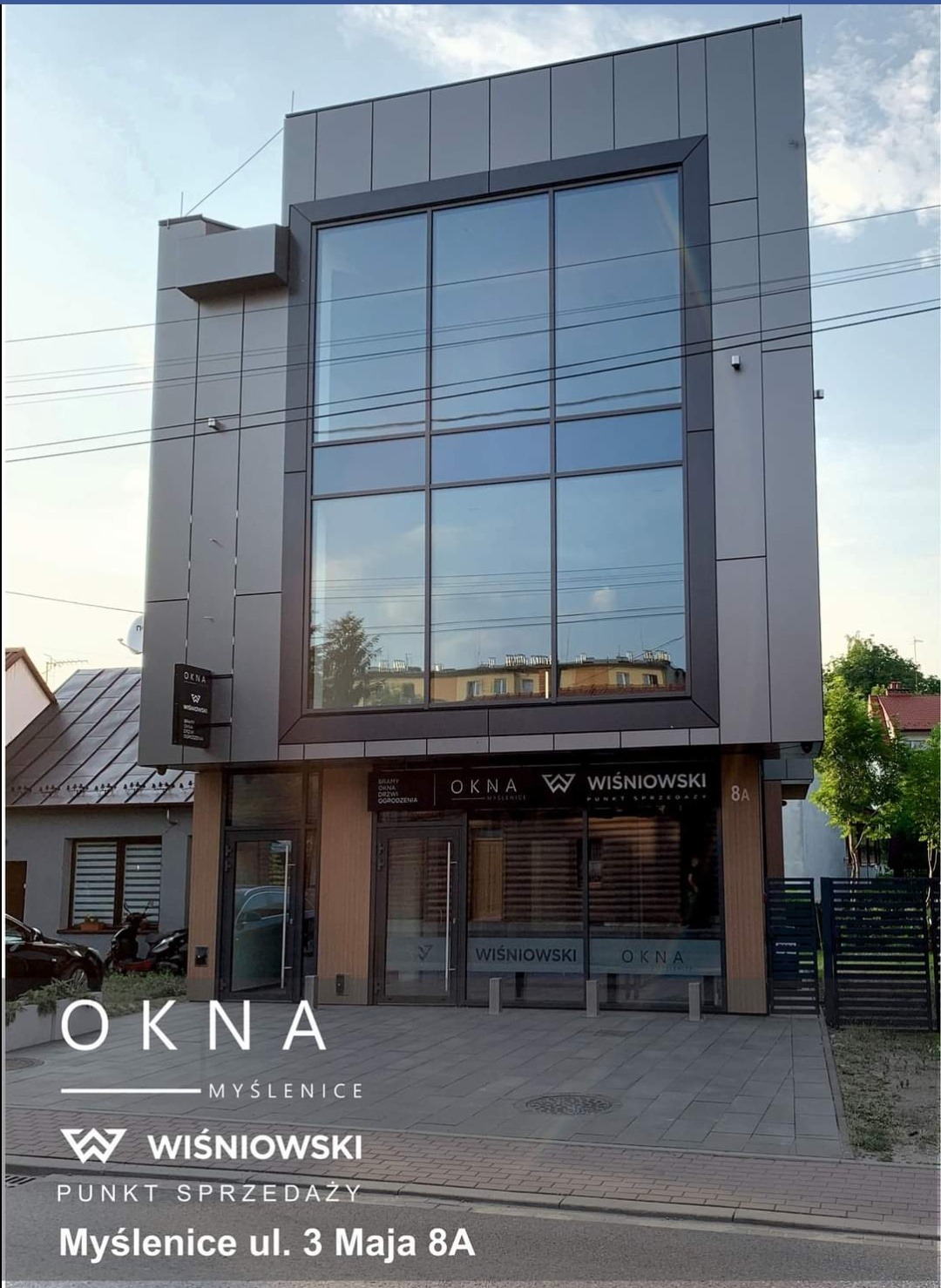 Nowoczesny budynek salonu sprzedaży stolarki okiennej i drzwiowej z dużym przeszkleniem, widok z zewnątrz, w tle zabudowa miejska.