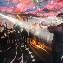 Iluzjonista w ciemnym garniturze, z mikrofonem nagłownym, wykonuje pokaz dla publiczności w sali z dekoracyjnym sufitem i wzorzystą podłogą.