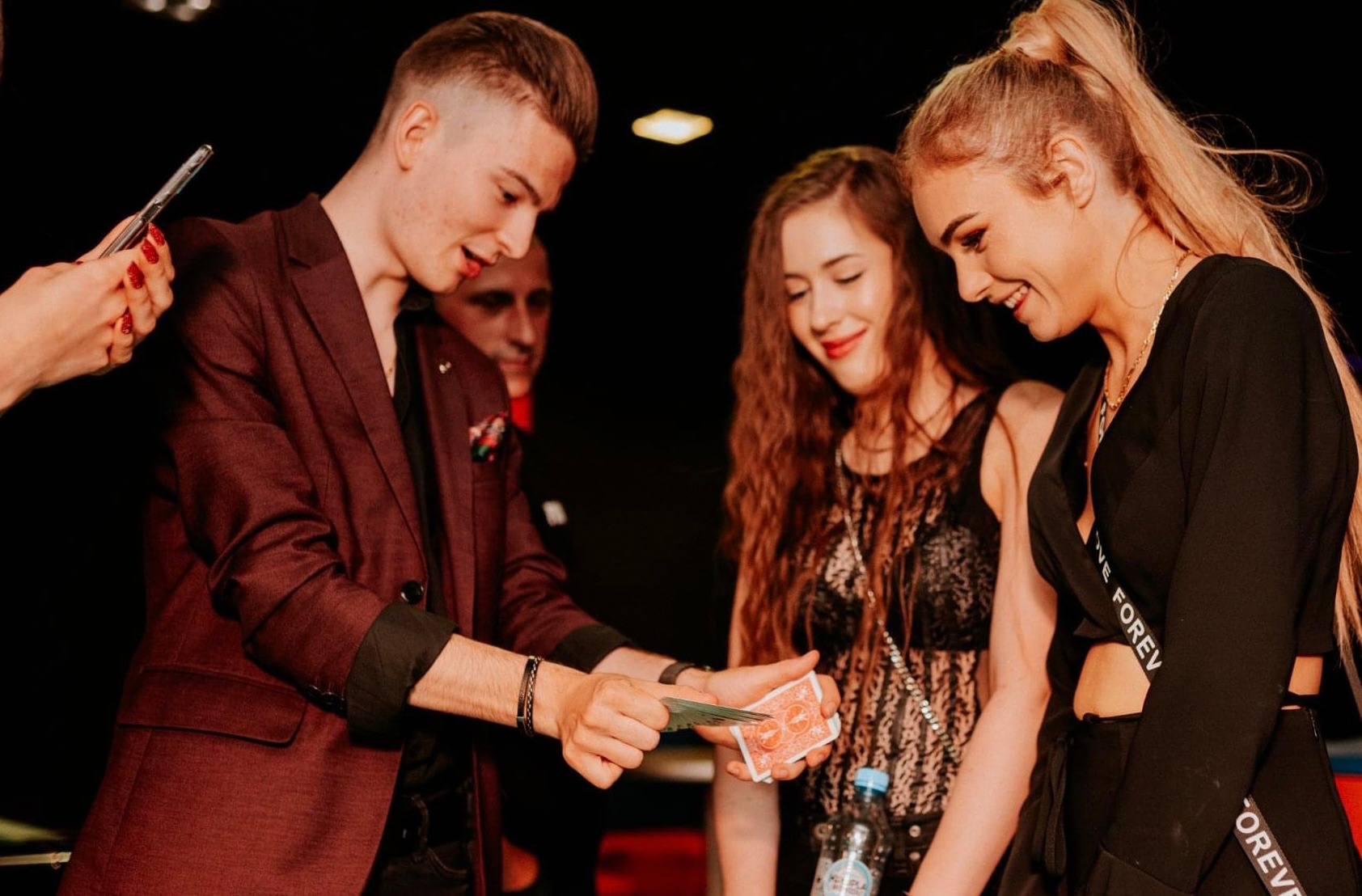 Młody mężczyzna w bordowym garniturze prezentuje trik karciany dwóm uśmiechniętym kobietom podczas występu, w tle widoczna twarz obserwatora.