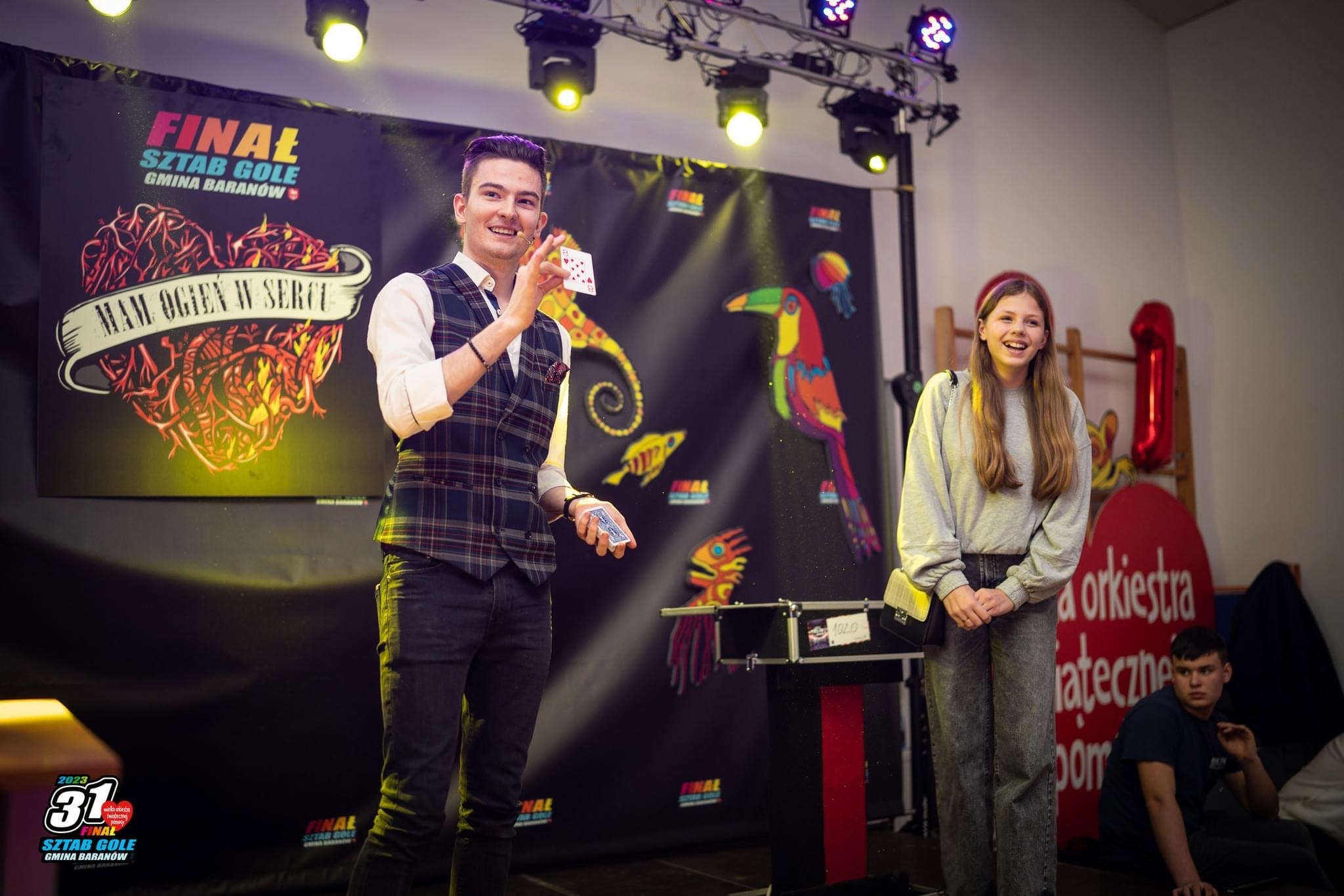 Uśmiechnięty iluzjonista w kraciastej kamizelce prezentuje kartę do gry na tle sceny z kolorowymi grafikami i publicznością.