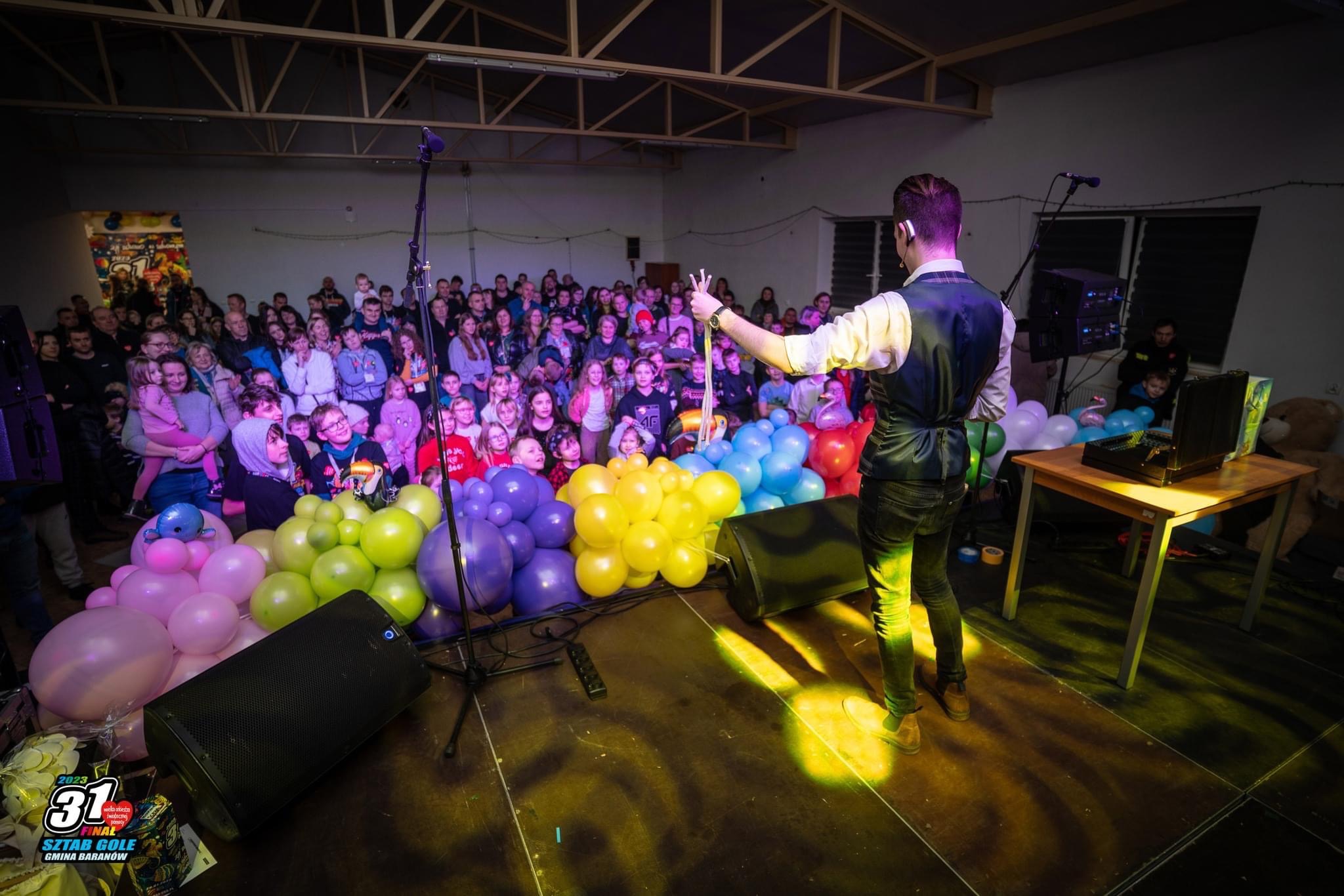 Iluzjonista w kamizelce prezentuje trik z sznurkami przed zgromadzoną publicznością, na scenie kolorowe balony i głośniki.