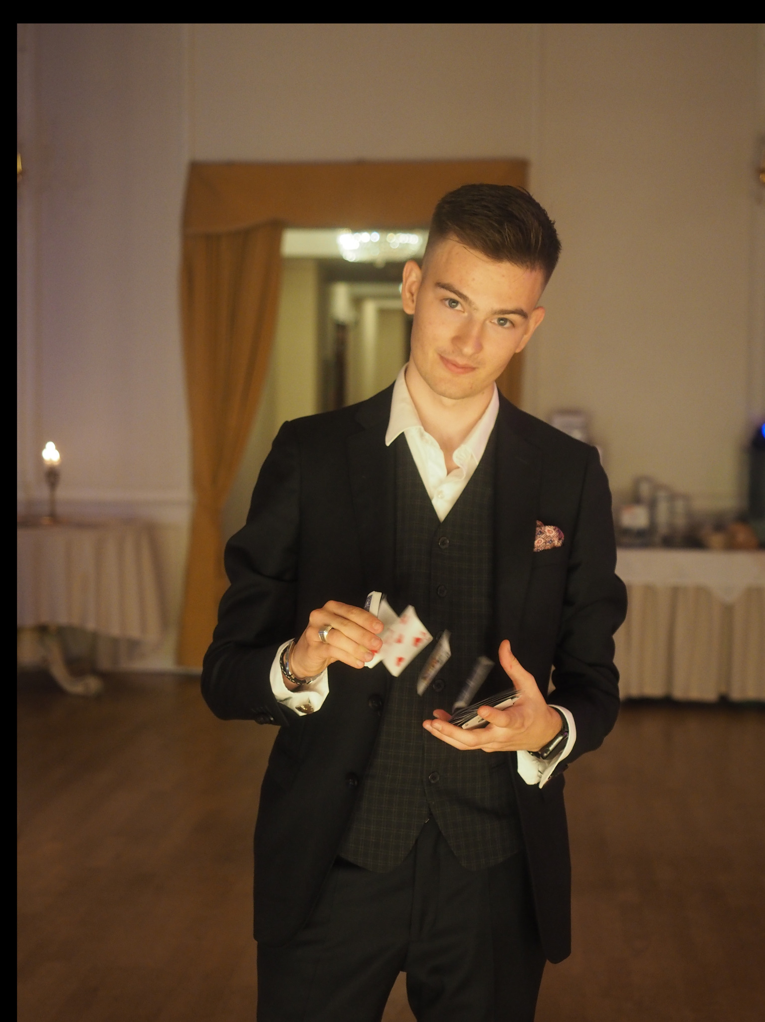 Młody mężczyzna w garniturze prezentuje lewitujące karty do gry na tle sali bankietowej z subtelnym oświetleniem.