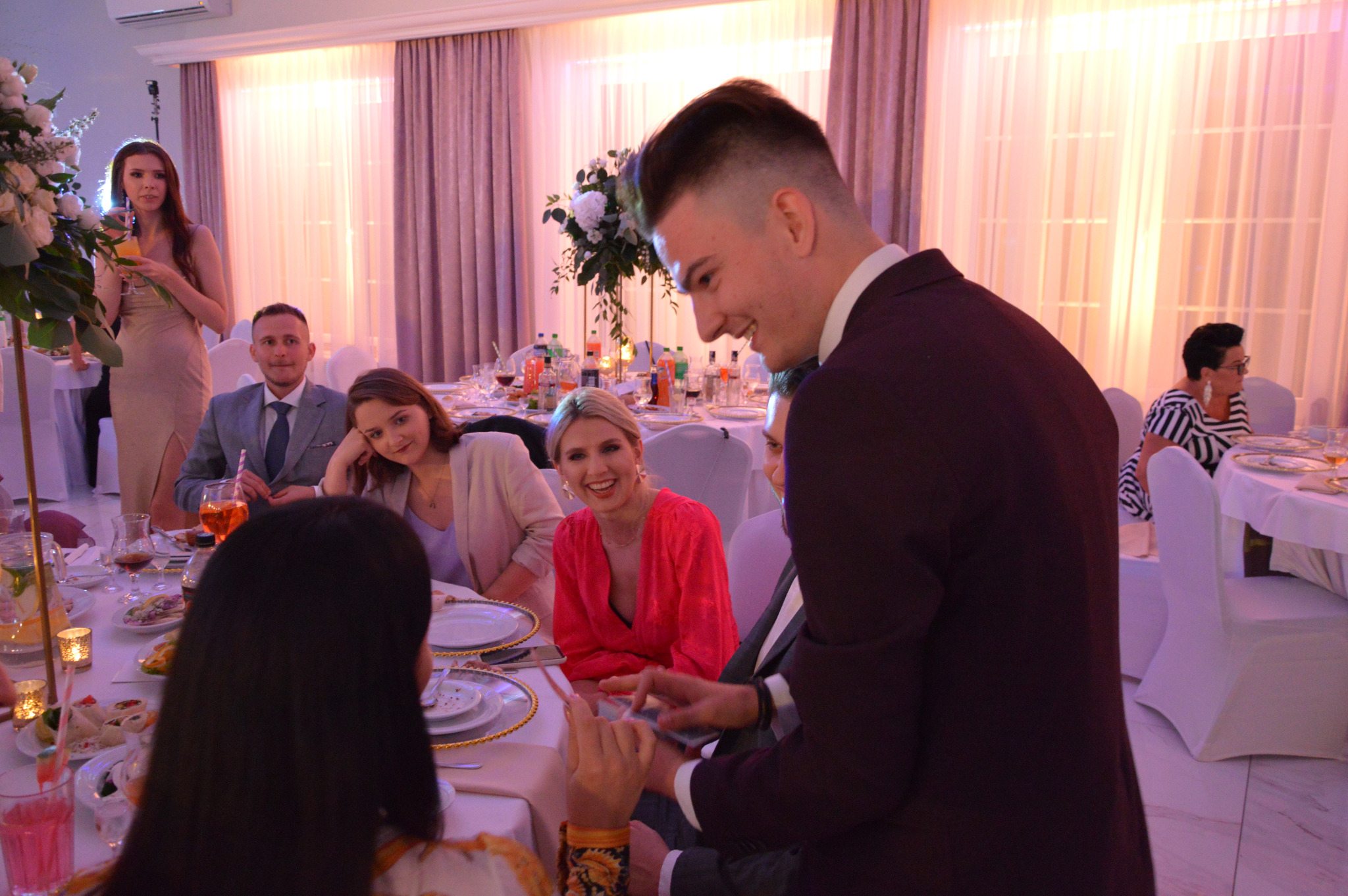 Uśmiechnięty iluzjonista w bordowej marynarce demonstruje trik z kartami przy stole gości podczas przyjęcia; w tle dekoracje kwiatowe i eleganckie oświetlenie sali.