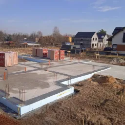 Zalany fundament pod dom jednorodzinny z izolacją termiczną, widoczne wystające pręty zbrojeniowe i palety z pustakami Porotherm w tle.