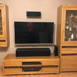 Zestaw mebli z jasnego drewna z ciemnymi akcentami: wisząca szafka z przeszklonymi drzwiami, szafka RTV pod telewizorem i wysoka szafka z przeszklonymi drzwiami, eksponująca szkło i butelki.