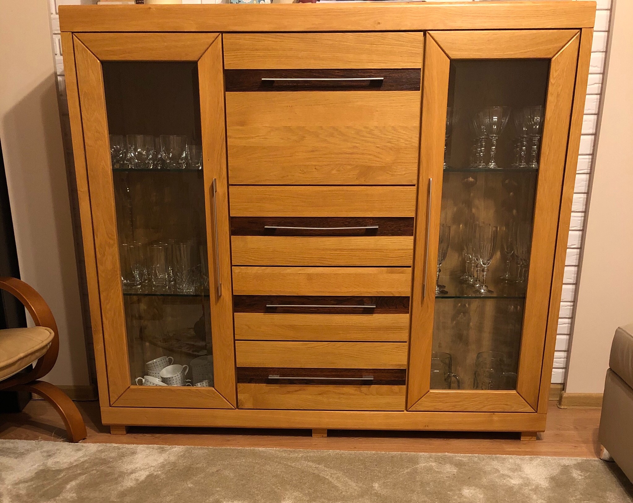 Drewniana witryna z przeszklonymi bokami, zawierająca szklanki i filiżanki, z czterema szufladami z ciemnymi wstawkami, stojąca na dywanie.