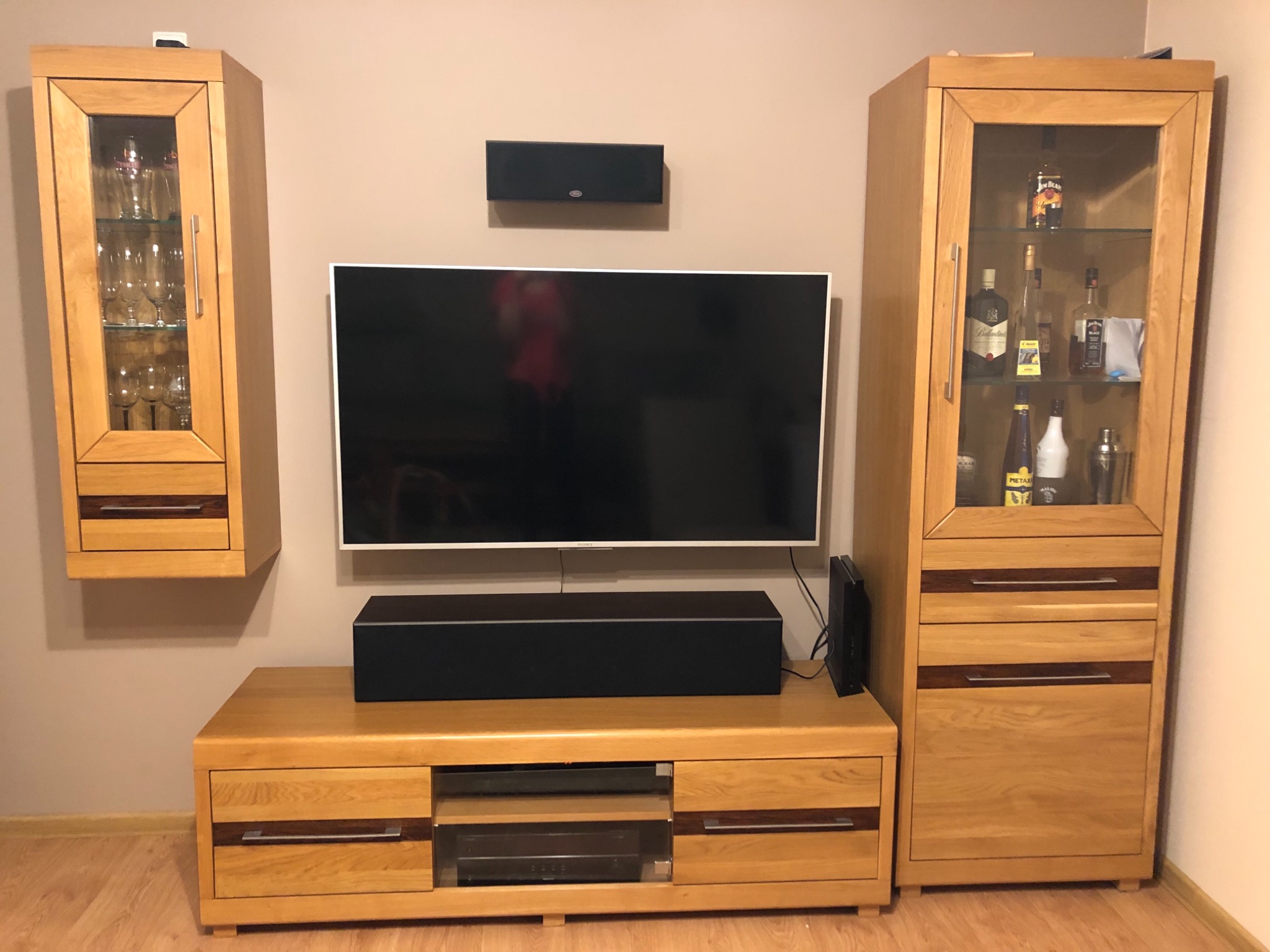 Zestaw mebli z jasnego drewna z ciemnymi akcentami: wisząca szafka z przeszklonymi drzwiami, szafka RTV pod telewizorem i wysoka szafka z przeszklonymi drzwiami, eksponująca szkło i butelki.