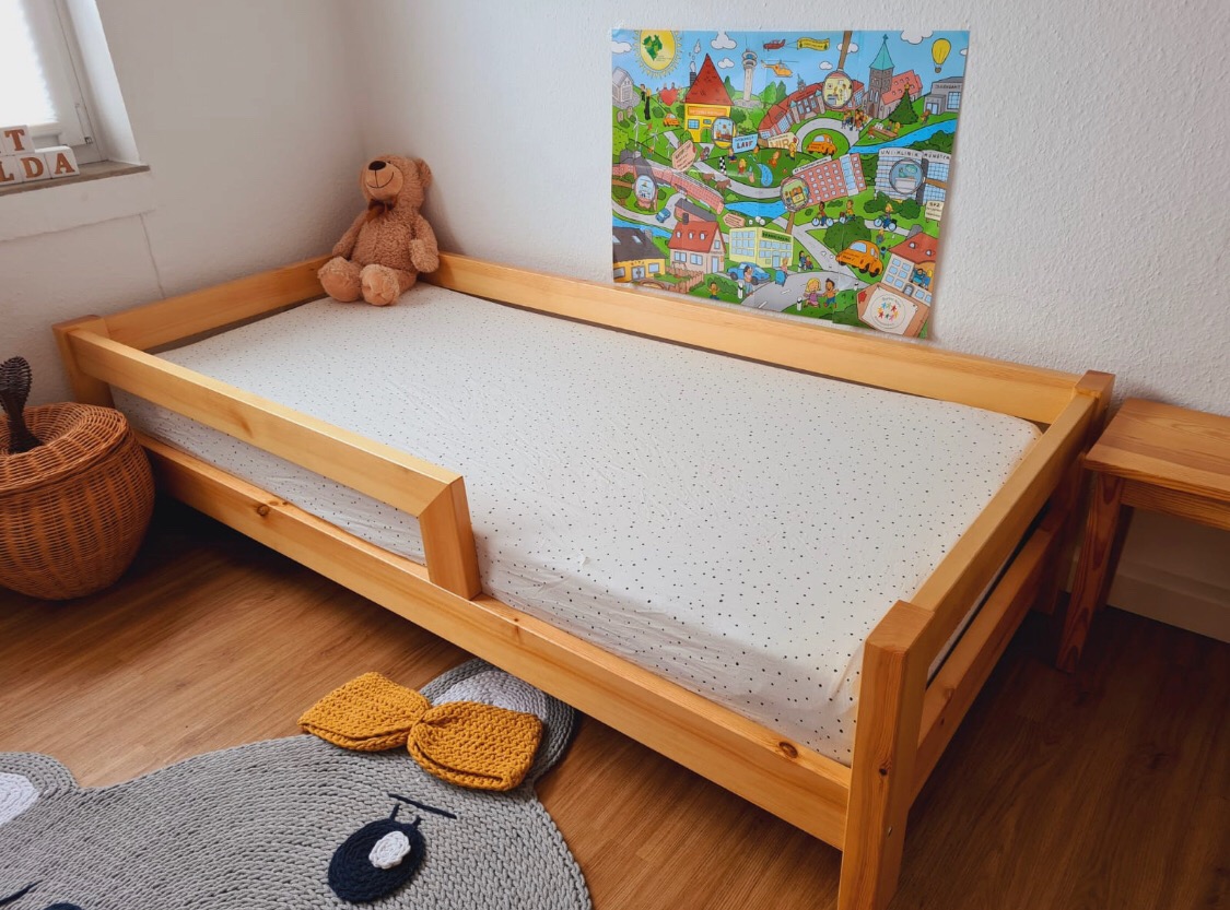 Drewniane łóżko dziecięce z barierką zabezpieczającą, pluszowy miś i kolorowa ilustracja na ścianie, uzupełnione plecionym koszem, dywanikiem w kształcie myszy i żółtymi kapciami.