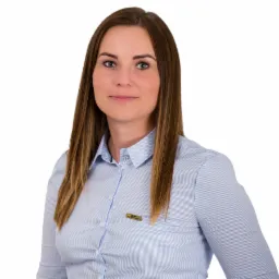 Portret kobiety w błękitnej koszuli z plakietką BHP, uśmiechająca się do kamery na białym tle.