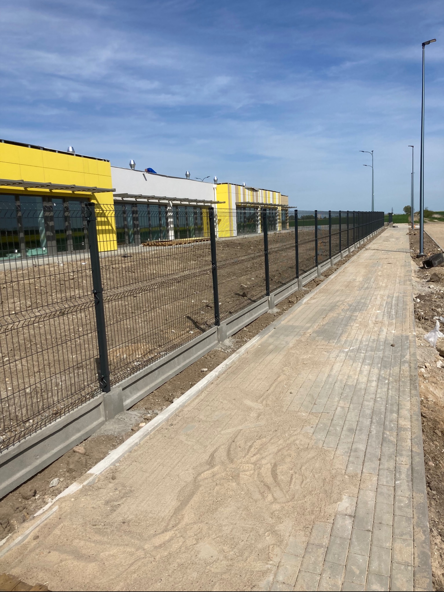 Długi, ciemnoszary system ogrodzeniowy z paneli na betonowej podmurówce, oddzielający budynek w kolorach żółto-białym od chodnika z kostki brukowej i terenu budowy w słoneczny dzień.