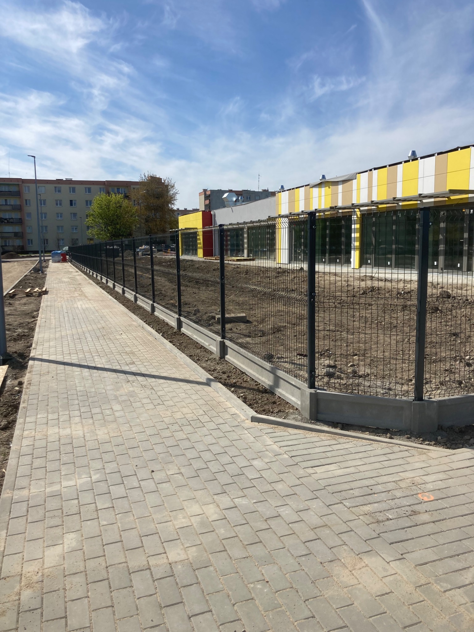 Ciemne, siatkowe ogrodzenie na betonowej podmurówce w Ciechanowie, oddzielające chodnik z kostki brukowej od terenu budowy z budynkiem w tle.