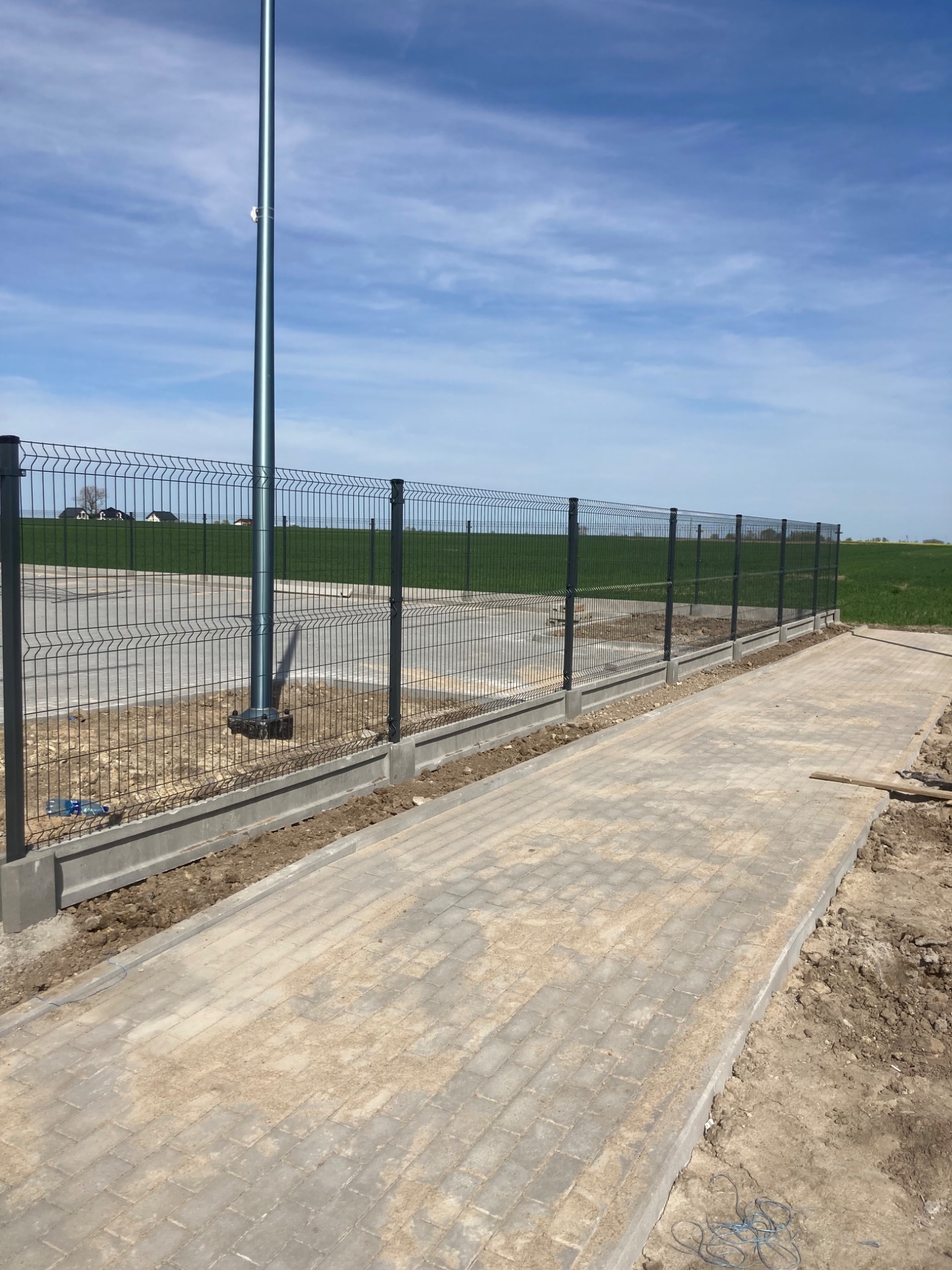 Nowo zamontowane szare ogrodzenie panelowe z podmurówką betonową, widoczne na tle kostki brukowej i słupa oświetleniowego, teren jeszcze niezagospodarowany.