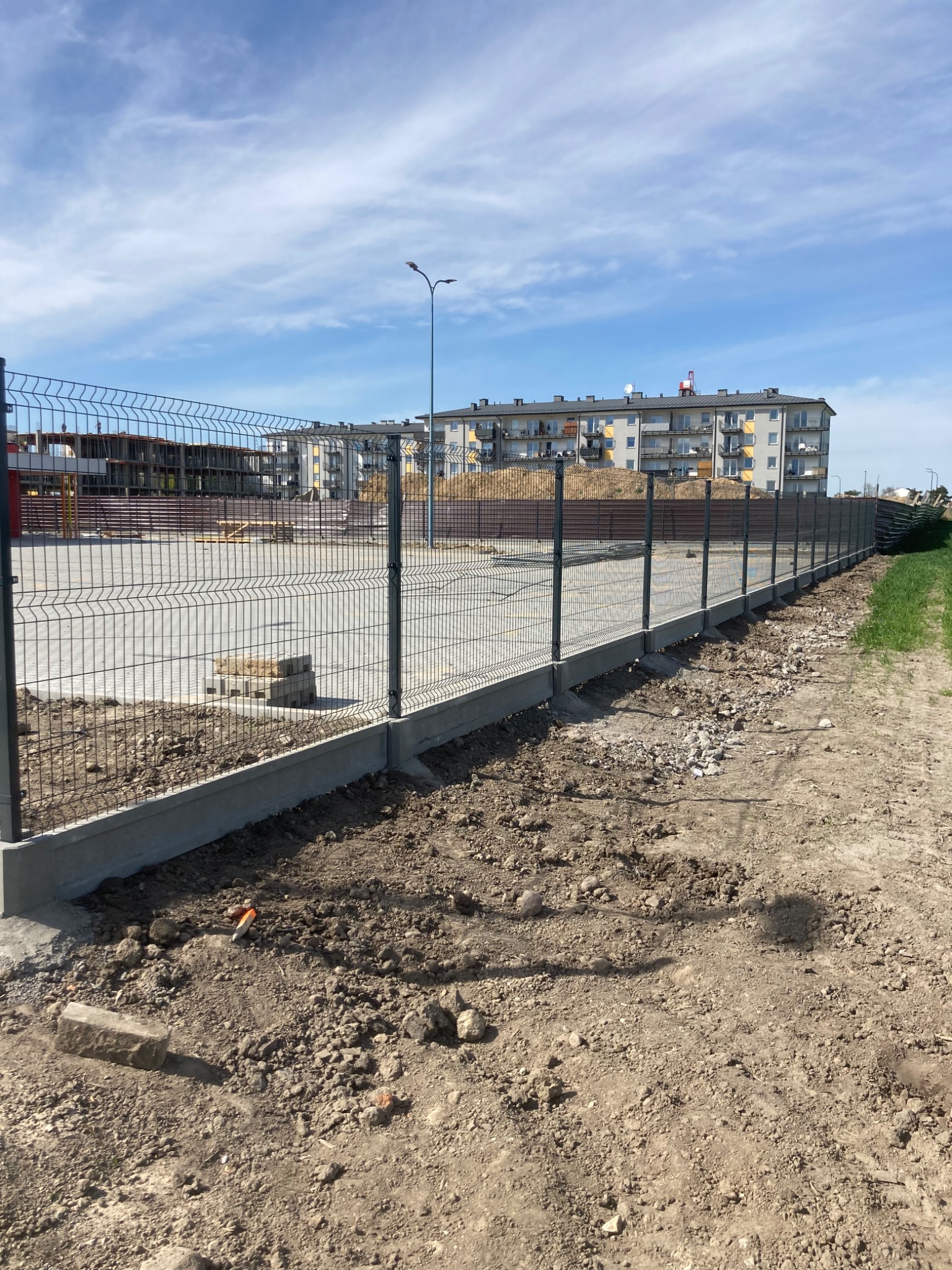 Nowo zainstalowane, grafitowe ogrodzenie panelowe z podmurówką betonową na terenie budowy, z widokiem na budynki mieszkalne w tle pod błękitnym niebem.