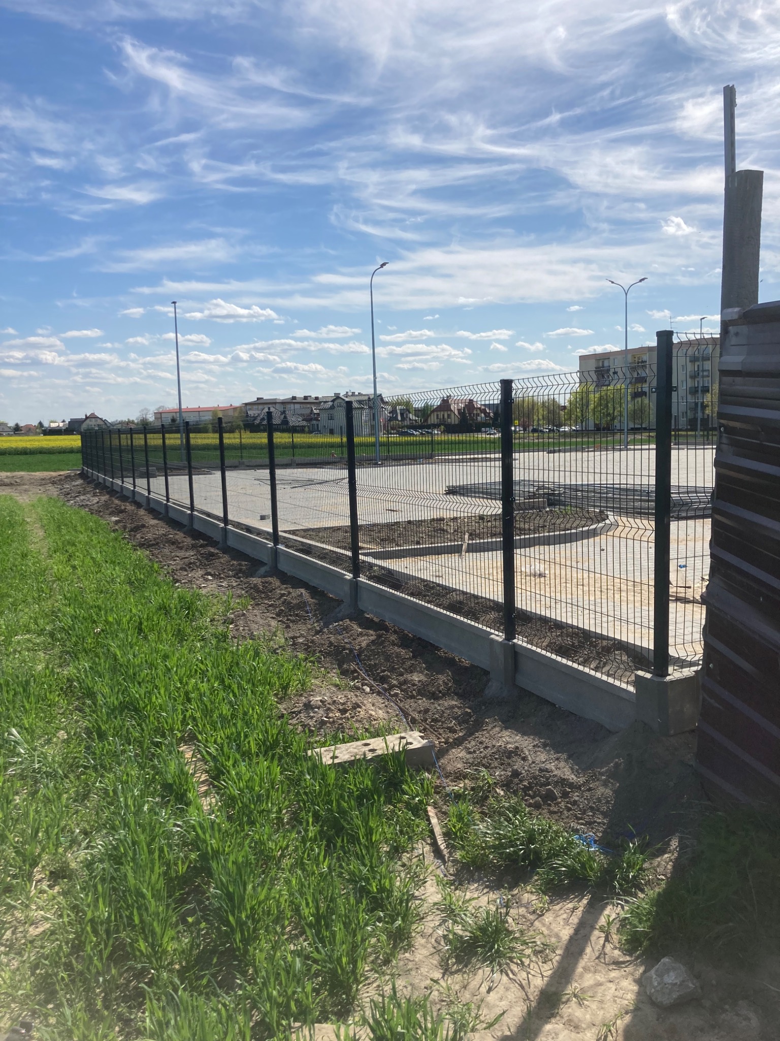 Nowo zainstalowane, ciemnozielone panele ogrodzeniowe na betonowej podmurówce, widok z boku na tle błękitnego nieba z chmurami i budynków w oddali, z widocznymi stertami paneli po prawej stronie.