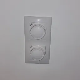 Biały, podwójny regulator temperatury na ścianie, z pokrętłami i symbolami fal, ustawiony w pozycji minimalnej.