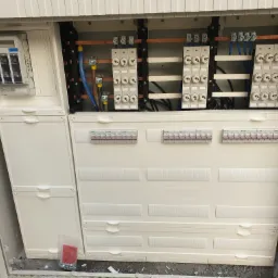 Otwarta skrzynka elektryczna Hager z bezpiecznikami, przewodami i wyłącznikami, widoczne elementy montażowe na szynach DIN, w tle szara ściana.