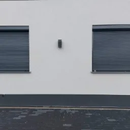 Dwa okna z całkowicie opuszczonymi, szarymi roletami zewnętrznymi na białej elewacji budynku, z widoczną kostką brukową w dolnej części kadru.