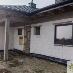 Surowy stan budynku w trakcie budowy z zamontowanym oknem w czarnej ramie, widoczne białe bloczki, brak elewacji, drewniane szalunki i podpory, oraz logo firmy w prawym dolnym rogu.