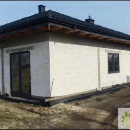 Dom w trakcie budowy, widoczne wstawione okna z ciemnymi ramami i drzwi balkonowe, ściany z białego bloczka, czarny dach, teren wokół domu niezagospodarowany, w prawym dolnym rogu logo firmy.